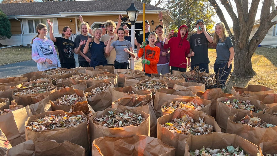 A large group of youth stand by bags of leaves that they raked.