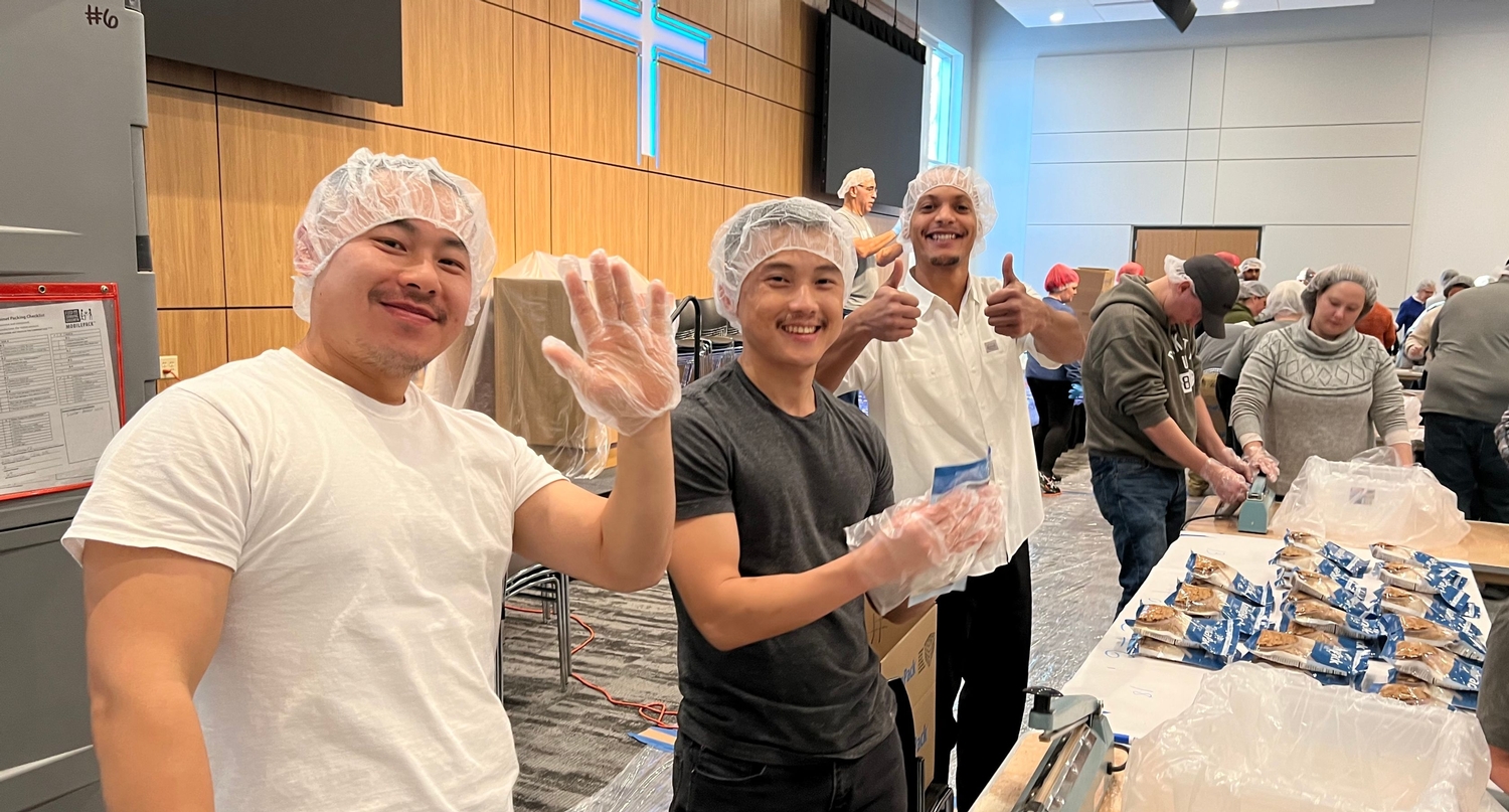Three men give a "thumbs up" while packing food for Feed My Starving Children